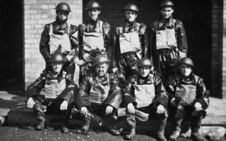 Photograph of eight men posed in two rows; each is wearing a steel helmet, overalls in a shiny substance, Wellington boots, a pack on his chest and a pair of gauntlets; they have been identified as Decontamination Squad, Easington Village