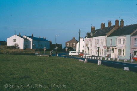 Photograph showing a view over a green with a road running along the right side of the green; at the end of the green another road can be seen running off to the right; at least two road signs instructing the driver to stop at the junction can be seen; a terrace of houses can be seen on the left , two buildings in the centre and a block of buildings, including The King's head and a shop, can be seen on the right; the picture has been identified as Cross Roads, Easington Village