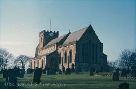 Photograph of the exterior of a church, showing the east end, a tower at the west end with crenellations and a clock, and part of the graveyard; it has been identified as St. Mary's Church, Easington Village