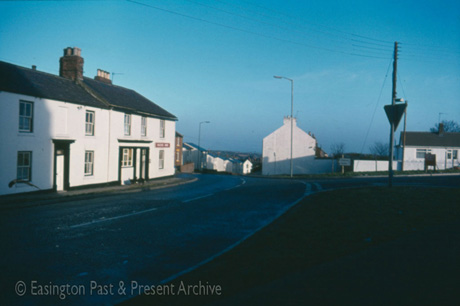 Photograph of a road in the foreground, running away from the camera over the brow of a hill; on the far side of the road are a house, painted white, with four windows and a door, adjoining a building with six windows and a doorway and a sign reading Masons' Arms; further buildings can be seen over the brow of, and at the top of,the hill; the buildings have been identified as being in Easington Village