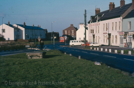 Photograph showing the same view as in easc0098, but from a point nearer to the crossroads; a triangle of green is in the foreground with roads running either side of it to a junction with a road running from the left and with one from the right; the same buildings can be seen, with the King's Head and the shop on the right being seen in more detail; two cars and a van are parked in front of the King's Head; the back of a bus can be seen on the road running away to the right; the photograph has been described as the Cross Roads in Easington Village