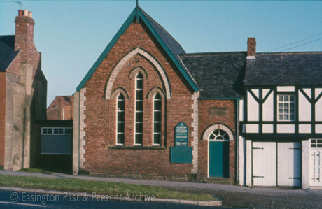 Photograph of the exterior of a brick chapel with three long narrow windows, adjoining a half-timbered building; there is a notice bord and a stone plaque on the chapel, neither of which can be read; the chapel has been identified as the Methodist Chapel in Easington Village
