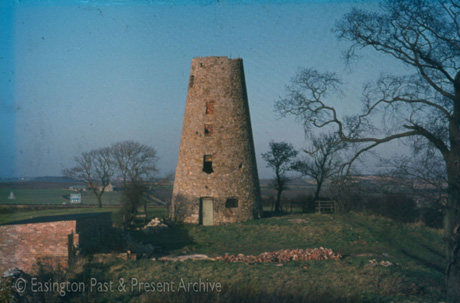Photograph of the circular building of a windmill with three widows one above the other and a doorway at the bottom; behind the mill are open fields and leafless trees; a blue road sign can be seen in the middle distance; the building has been identified as Easington village Mill