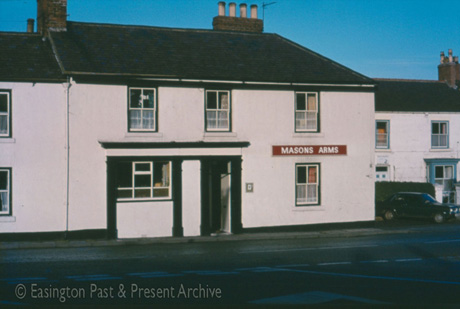Photograph, taken from across a road, close-up, of the exterior of the Masons' Arms; the building is painted white, is two-storeyed and has four sash windows; a fourth, 1970s, window is on the left of the doorway and is flanked, as is the doorway, by two black columns; the building has been identified as being in Easington village