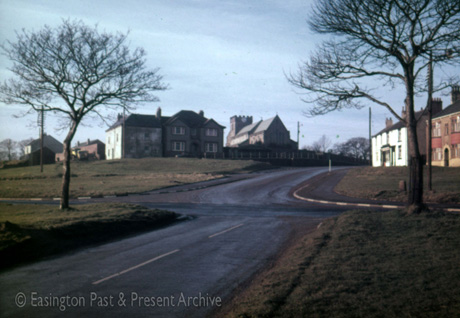 Photograph, looking uphill, past green open space, along a road towards the east end of the church and a the front of a large house; another road crosses that running uphill in the middle ground; the photograph has been described as Village Green, Easington Village