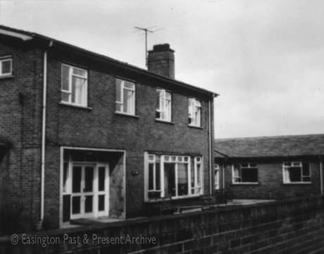 Photograph of the exterior of brick house, of the 1960s or 1970s, with four windows on the first floor and a large window and a large doorway on the ground floor; a single-storey wing appears to form two sides of a square at the right of the building, which has been identified as Essyn House, Easington Village
