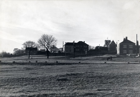 Photograph showing, in the foreground, an open expanse of grass with substantial houses on its far edge; the end of a church with a crenellated tower and a clock can also be seen; a horse is grazing on the grass; the scene has been identified as Easington Village