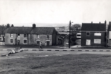 Photograph showing, in the foreground, a piece of grass and a road with a roadworks sign and barrier; beyond the road are three cottages possibly built in the nineteenth century and a small house built after the Second World War, which is bricked up; beyond these buildings low buildings of the post-war era can be seen and beyond those a view into the far distance over a plain; the photograph has ben identified as Easington Village