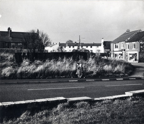 Photograph showing a road running across the photograph with two women walking on the far pavement; beyond them is an overgrown verge, the wall and roof of a house; beyond the wall, the upper storeys of a terrace of houses, a road sign and the fronts of shops can be seen; none of the details of these can be seen as they are too far from the camera; the photograph has been identified as Easington Village