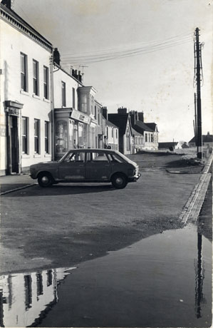 Photograph showing, on the right, the facades of a pre-twentieth century house with a pillared doorway, the front of two shops and other buildings of some age forming a continuous facade; in front of the first building a car is parked; no details of the buildings or the car can be discerned; the photograph has been identified as Easington Village
