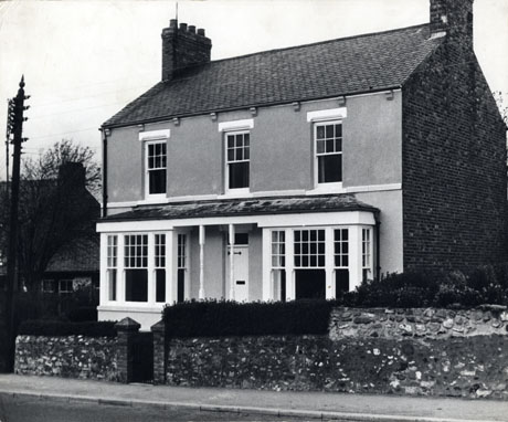 Photograph of the facade of a house with two square bay windows on the ground floor and three windows on the first floor; the door of the house is protected by a porch continuous with the bays; there is a small front garden behind a low wall which runs along the pavement; the house has been identified as being in Easington Village