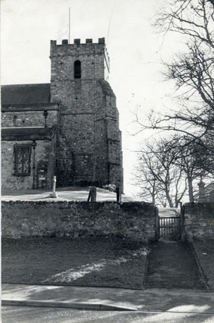 Photograph showing the wall of the churchyard, three gravestones and the exterior of the crenellated tower and nave of a church; a flag of St. George is flying on the tower; the church has been identified as Easington Village
