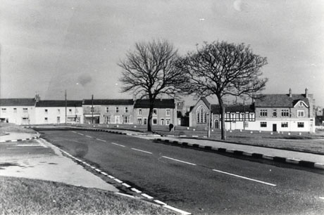 Photograph showing a road running away from the camera towards a row of four houses at the edge of a green; on the right of the houses is a chapel, which is attached to a half timbered building which is attached to a large building which may be a public house; the photograph has been identified as depicting Easington Village