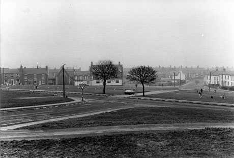 Photograph showing a road running across the green from right to left of the photograph with another road crossing it in the other direction; the houses adjoining the chapel can be seen on the left of the photograph and houses on the distance away from the green can be seen in the right of the photograph; the photograph has been identified as being in Easington Village
