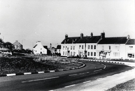 Photograph showing a road running away from the camera to the right; on the right of the road is a large building with the words King's Head on it; on its right are two buildings with unidentifiable shops in them; on its left are the ends of three buildings; on the left of the picture is grass on the left of the road; the scene has been identified as being in Easington Village