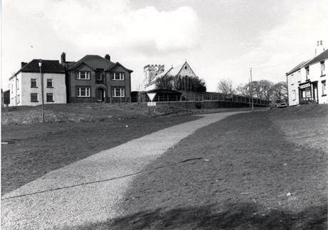 Photograph showing a surfaced road running through grass towards a church, the top of the end and tower of which can be seen above a wall and trees; on the left of the church are two large houses; on the right are two houses in one of which is an identifiable shop; the scene has been identified as being in Easington Village