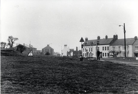 Photograph showing, across grass, the King's Head on the right and another two houses attached to it on its right; on its left, the ends of three houses and the front of one house can be seen; a sign with an exclamation mark on it is on the green, which takes up most of the photograph; a man is standing at the bus stop on the right of the green; the scene has been identified as being in Easington Village