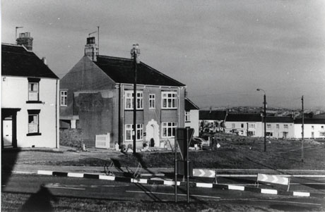 Photograph showing a road in the foreground with a barrier directing traffic down one lane of the road; in the background are the end of a house on the left and the front and side of a detached house in the centre of the photograph; in the distance on the right are terraced houses and indistinct buildings in the far distance; the photograph has been identified as Easington Village
