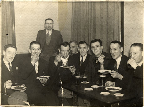 Photograph of eight men, wearing suits, sitting in a room round a small table on which there are four glasses of beer; four of the men are holding plates of peas; another man is standing behind those sitting; they have been identified as being in Easington Village