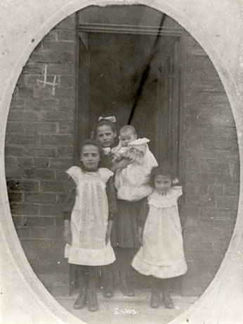 Photograph of four girls in the doorway of a brick house; two girls aged approximately four and six years are wearing white pinafores and standing in front of the doorway; an older girl aged approximately ten years is wearing a dress and a ribbon in her hair and standing in the doorway behind the other girls; she is holding a baby in her arms; the children have been identified as Allinson children