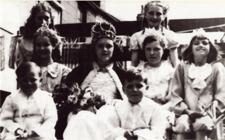Photograph showing six adolescent girls dressed as a queen and her acolytes with two small boys, presumably the queen's pages; the queen is wearing a crown and a cloak and carrying flowers; the upper storey of a house can be seen behind the group and Union Jacks can also be seen and one of the girls is carrying a Union Jack; the photograph is described as Haswell Queen-I.O.G.T.; I.O.G.T. refers to the Order of the Good Templars it must be assumed