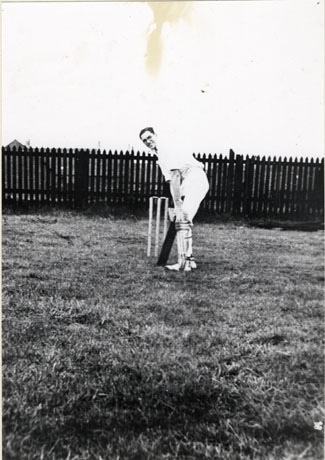 Photograph of a man identified as Jack Dormand wearing cricket whites and standing in front of cricket stumps ready to bat; he is also wearing glasses; behind him is a fence and behind that what appears to be the roof of a building; the photograph is described asJack Dormand, Haswell Cricket Club