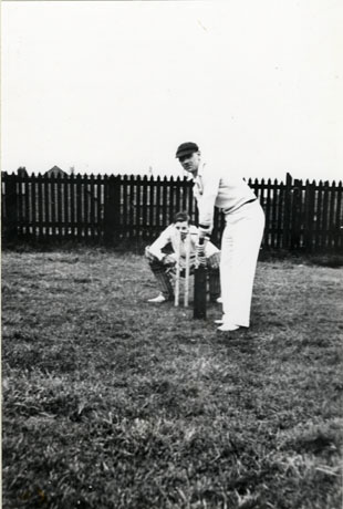 Photograph of a man in cricket whites in front of cricket stumps preparing to bat; behind him is another man squatting and acting as wicket keeper; the photograph has been taken an the same place as hasw0004 and a fence is immediately behind the men; the batsman has been identified originally as Steve Barry and later as Arthur Wright; the wicket keeper has been identified as Wilf Smith