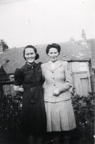 Photograph of two women in front of bushes, a fence and a building which appears to be an outhouse; one woman is wearing a suit with a long jacket and box-pleated skirt; the other woman is wearing a short-sleeved dress; the women have been identified as Jenny and Marion Featonby, 47, Old Front Street, Haswell