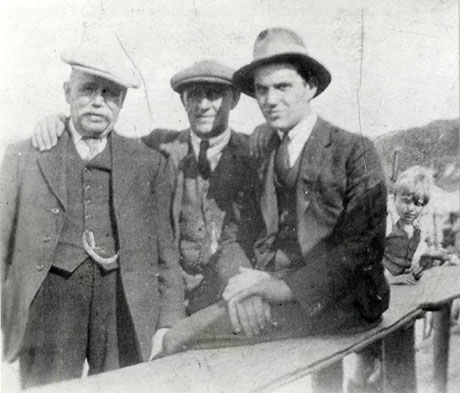 Photograph of an elderly man with a moustache wearing a suit, tie, cap, and watch chain, standing with another man who is behind him and resting his right hand on the elderly man's shoulder; this man who is wearing a suit, tie, and cap is also standing and resting his left hand on the shoulder of a third, younger, man who is perched on a parapet; the younger man is also wearing a suit and tie and a Trilby hat; behind the three men a small boy can be seen; the men have been identified as Eddie Taylor, Philip Hunter Featonby, and friend