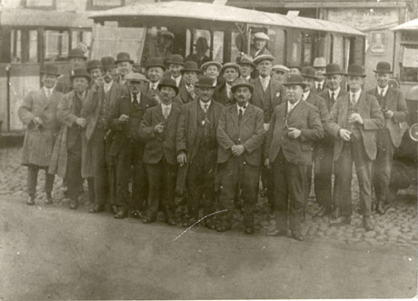 Photograph of a group of twenty five men in suits, bowler hats, Trilby hats and caps standing in front of a motor bus; part of the front of a car can be seen at the right-hand side of the picture; the photograph has been identified as Businessmen's Outing