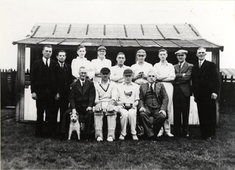 Photograph of thirteen members of Haswell Cricket Club Team, posed in front of a wooden structure with a verandah, possibly the pavilion; seven of the members are wearing cricket whites; the others are dressed conventionally in suits and ties; one man at the left end of the front row is holding the collar of a small terrier dog; a man second from the right on the front row is holding a trophy