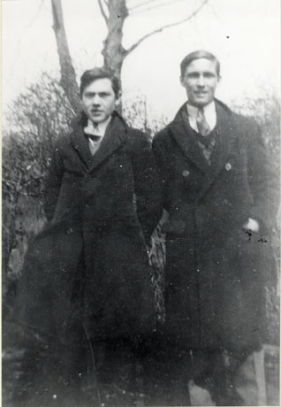 Photograph, full- length, of two young men, dressed in overcoats and collars and ties, and posed against a hedge and tree; they have been identified as Norman Parkin and Tom Featonby