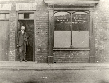 Photograph of the exterior of the First Insurance Office, Haswell showing a doorway and shop window in a brick wall; on the window are written the words Fire, Life, Accident, Burglary, Motors & General Insurance; above the window is an illegible series of words; in the doorway a man dressed in a suit and tie is standing