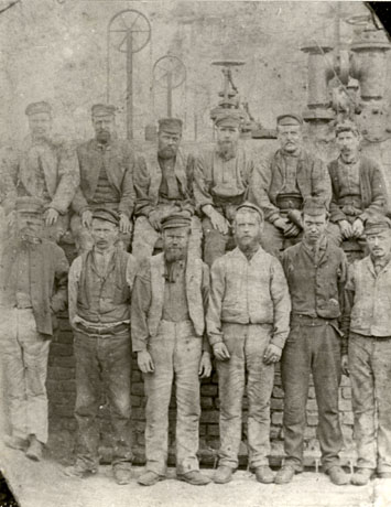Photograph of twelve men in working clothes identified as miners at Haswell Colliery; the miners, who are posed against a very faintly seen wall with pipes above it, are all wearing caps with a high crown and small brim