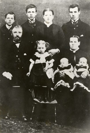 Photograph of family of two adults, three young men, one young woman, one young girl of approximately four years of age, and infant twins; the family is posed against a indistinct background, possibly in a photographer's studio, and the small child is standing on a Windsor chair