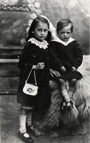 Photograph of a small girl of approximately six years of age standing with a small boy of approximately four years of age; the girl is wearing a dress with a large lace collar, buckled shoes and light- coloured socks and a ring on her right hand; she is also carrying a handbag;the boy is sitting on couch covered with long-haired material and is wearing a sailor suit, light-coloured socks and buckled shoes; the background is indistinct and may be a photographer's studio; they have been identified as Eleanor Carr Allinson and her brother