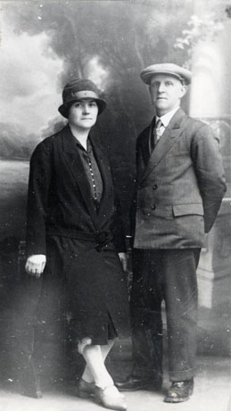 Photograph, full-length, of a man and a woman taken in a photographer's studio; the woman is wearing a felt hat with a brim low down on her forehead, a coat fastened on the hips, gloves, and pointed buckled shoes; the man is wearing a flat cap, a double-breasted suit and brogues; they have been identified as Philip Featonby and Liza Ann Featonby