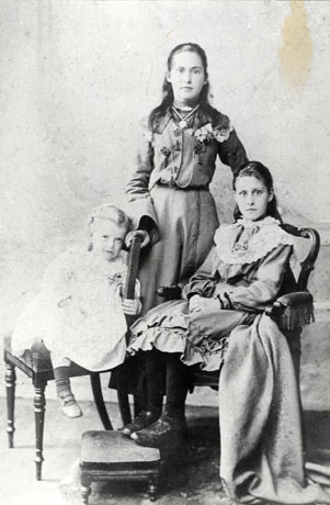 Photograph, full-length, of a girl of approximately fifteen years, standing behind a girl of approximately thirteen years, sitting in a chair with arms, and behind a girl of approximately four years sitting on an upright chair; the two older girls are wearing dresses with lace, frills and embroidery on them; the dress of the child is indistinct; the photograph has been taken in an photographer's studio; the girls have been identified as Jenny Heatherington and sisters