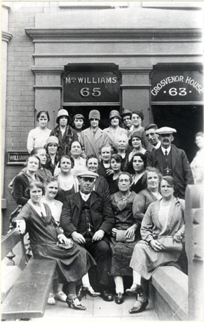 Photograph of a group of twenty two women and four men outside the doorways of two buildings, above which are written the following: Mrs. Williams 65 and Grosvenor House 63; the group has been identified as Coach Party from Haswell Visiting Blackpool