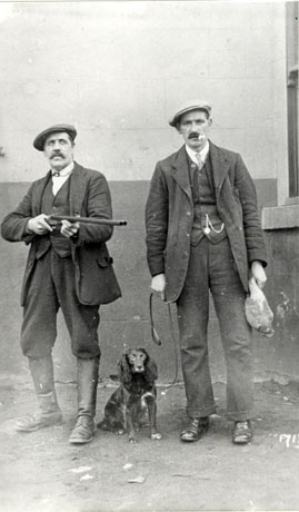 Photograph, full-length, of two men posed against the wall of a building; one man, who is dressed in a cap, a long jacket, and gaiters, is holding a gun; the second man is holding the lead of a Spaniel in his right hand and a dead bird in his left; he is dressed in a suit and a flat cap; the men have been identified as Henry Allinson and Jack Hornsby