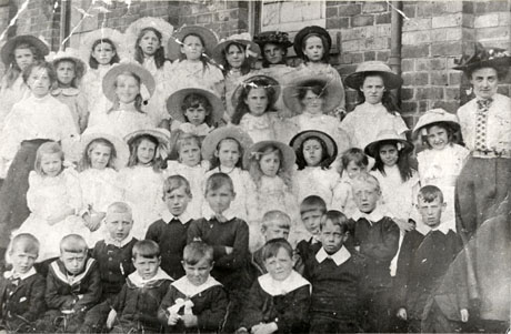 Photograph of twenty three girls and thirteen boys posed against a brick wall with two women on either side of them; the girls are dressed in light-coloured clothes and are all wearing hats; the boys are dressed in suits with Eton collars; the group has been identified as Haswell Infant and Junior School, Class 2A, 1907