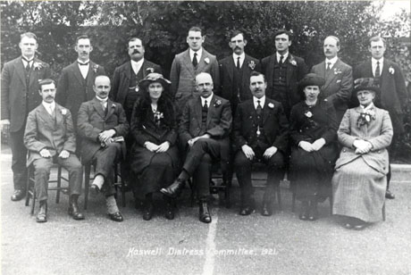 Photograph entitled Haswell Distress Committee, showing a group of twelve men and three women posed against trees and on tarmac; the men, who are wearing suits, all have a button- hole in their lapels; the women, who are also formally dressed in jackets and skirts are also wearing flowers