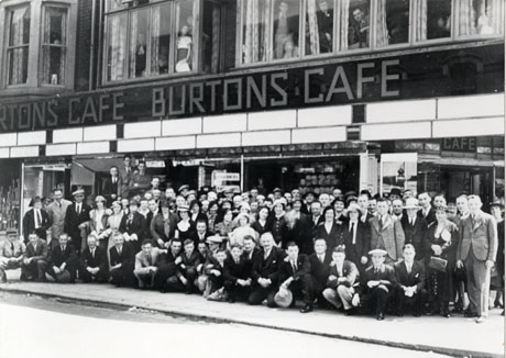 Photograph of approximately eighty men and women posed on the pavement outside a building bearing the words Burtons Cafe;part of the windows of the cafe can be seen at ground-floor level behind the group; people can be seen at the windows on the first floor looking out at the group; the photograph has been identified as depicting an outing of the employees of the Northern Sabulite works to Blackpool