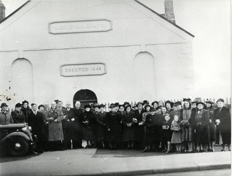Photograph of approximately thirty five people outside a chapel; the tops of railings can be seen behind them and the wall of the West end of the chapel on which are written the words Wesleyan Chapel and Erected 1849; at the left-hand side of the photograph, the bonnet of a car can be seen; a clergyman can be seen in the middle of the group; the photograph has been identified as being taken on the occasion of the chapel's centenary