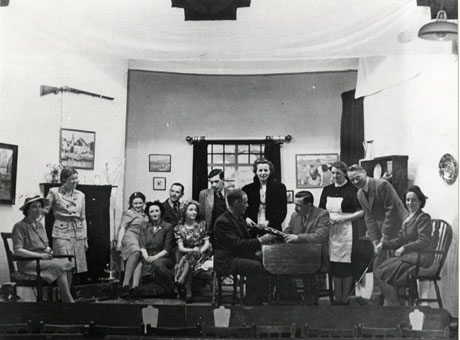 Photograph of the cast of a production of Quiet Wedding on stage and in costume; the set appears to be that of the interior of a middle-class house and the costumes appear to reflect the dress of the nineteen forties; thirteen people can be seen on stage, including a maid and a golfer; the photograph has been identified as depicting amateur dramatics
