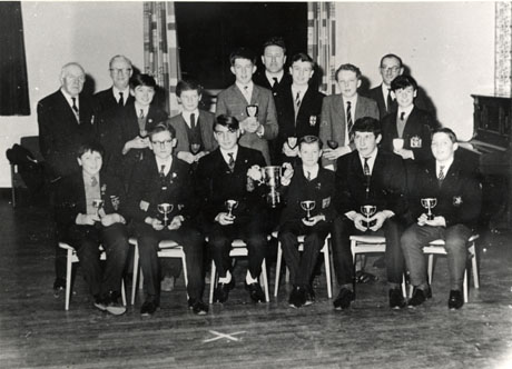 Photograph of twelve schoolboys, with four middle-aged men, in what appears to a school hall, with an upright piano on the right-hand side and a window behind the group; each of the boys is holding a small cup and two boys on the front row are holding a large cup between them; all but three of the boys are wearing school blazers and the badges on the pockets seem to vary; the photograph is described as Final of County Road Safety Quiz, 1965