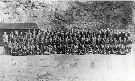 Photograph of a group of approximately one hundred and ten members of the staff of The Northern Sabulite Explosives factory, posed against the walls of a quarry; the photograph is taken from a distance but the faces of some of the members of staff can be discerned