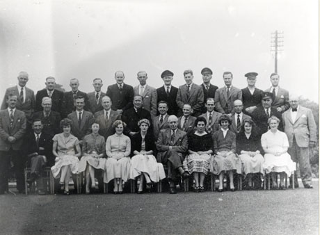 Photograph of a group of twenty five men and eight women posed in the open air ; the women are sitting on chairs on the front row and all except two men are standing round them; four of the men are wearing a uniform and peaked cap; all the other men are dressed in jackets and ties; the women are dressed in full skirts and cardigans; the photograph has been identified as depicting I.C.I. Office Staff, 1957