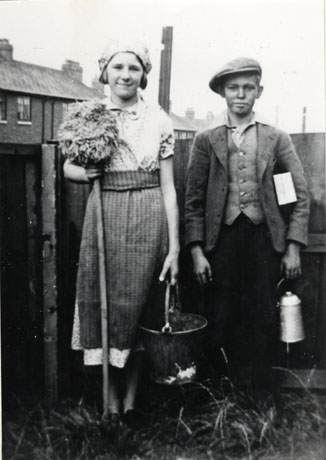 Photograph of a girl aged approximately fourteen years and a boy aged approximately twelve standing in the garden of a house; the upper storey and roof of a house can be seen in the distance and grass can be seen at their feet; the girl is wearing a dress and apron and has a scarf round her head; she is carrying a mop and a bucket; the boy is wearing a flat cap, a jacket and waistcoat; he is carrying a lunch box under his arm and has a can for tea or other liquid refreshment in his hand; the children are identified as Salkeld Children, Haswell