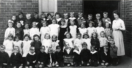 Photograph of forty nine children, aged approximately five years, posed outside a brick building with their female teacher; a child on the front row is holding a notice reading: Haswell Infant School Class 4 1919